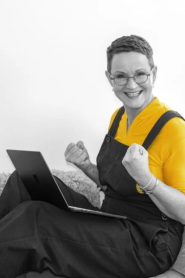 Imke Koop sitzt mit ihrem Laptop im Schoß auf einer Couch und ballt ihre Hände zu siegessicheren Fäusten als Symbolbild zu warum sie sich für Voice Technology begeistert. Das Bild ist in schwarz-weiß, lediglich ihr Shirt leuchtet in strahlendem Gelb.